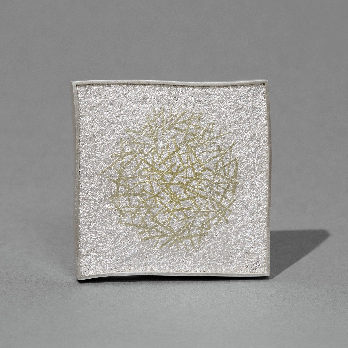 Square textured white silver brooch with gold abstract design on a gray background